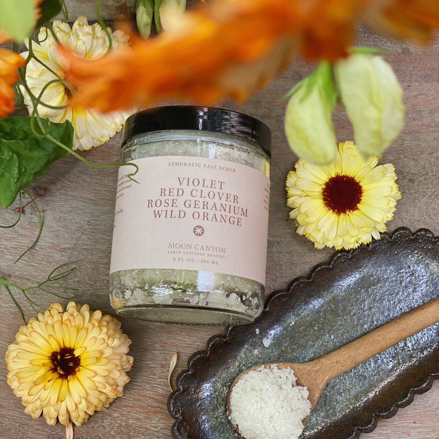 A jar of Moon Canyon Healing's Violet, Red Clover, Rose Geranium & Wild Orange Lymphatic Salt Scrub is placed on a wooden surface. Next to it, a small decorative dish holds a wooden spoon with some of the detoxifying scrub. Surrounding the items are yellow and orange flowers, enhancing the scene’s emphasis on skin health.

