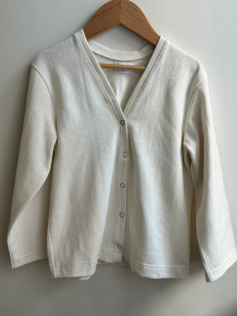The TAKE CARE TOP hemp by cabane childrenswear, an ethically handmade, long-sleeved off-white cardigan with a V-neck and button-down front, hangs on a wooden hanger against a light wall.