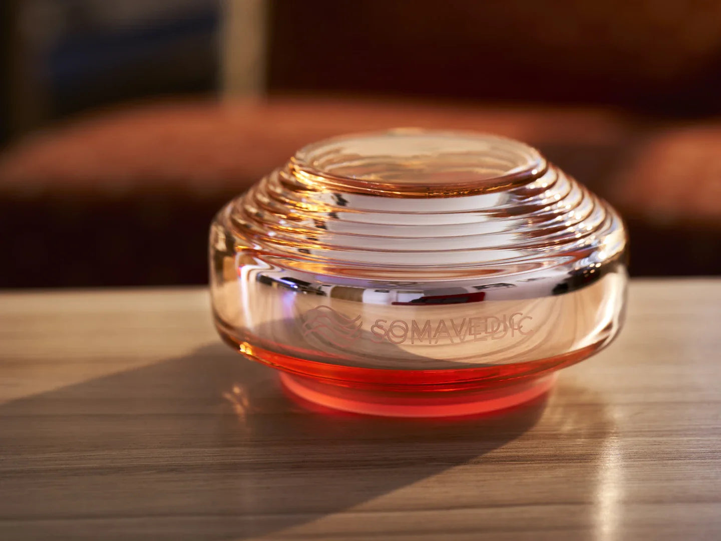 The Somavedic USA Rosalin, a round glass device with amber and red tints and horizontal ridges, sits on a wooden surface. Its hues evoke red light therapy benefits. "Somavedic" is etched on its side; the background is softly blurred.