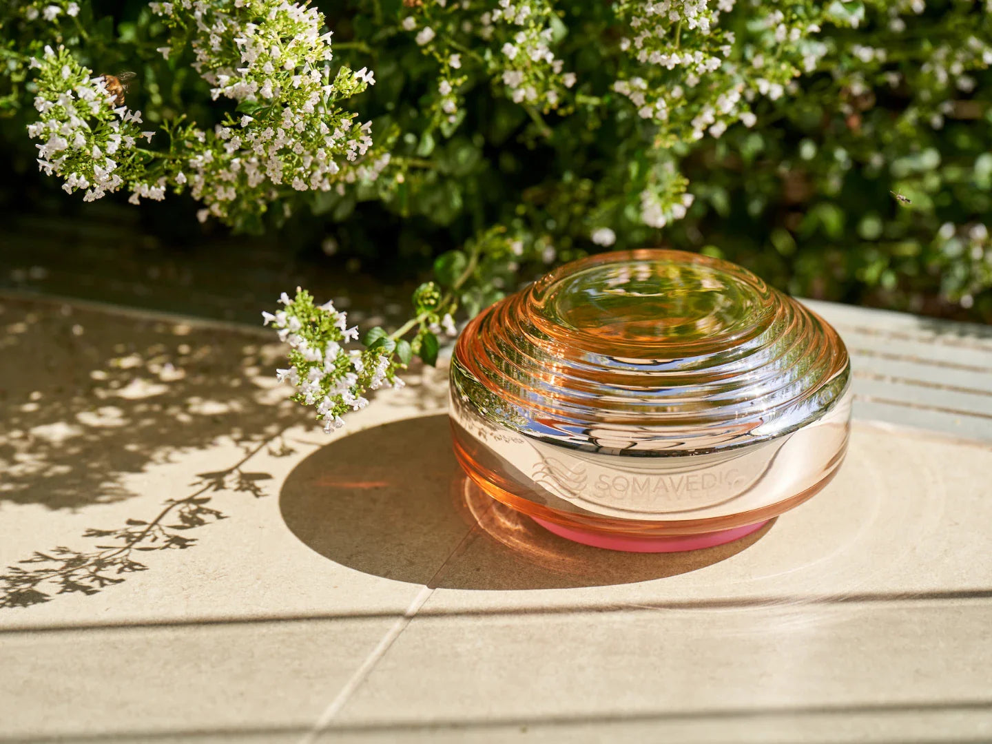 A round, glass, dome-shaped object labeled "Rosalin" by Somavedic USA rests on a sunlit tiled surface near green foliage with white flowers, casting a soft shadow—evoking gentle circadian rhythm support in a tranquil setting.