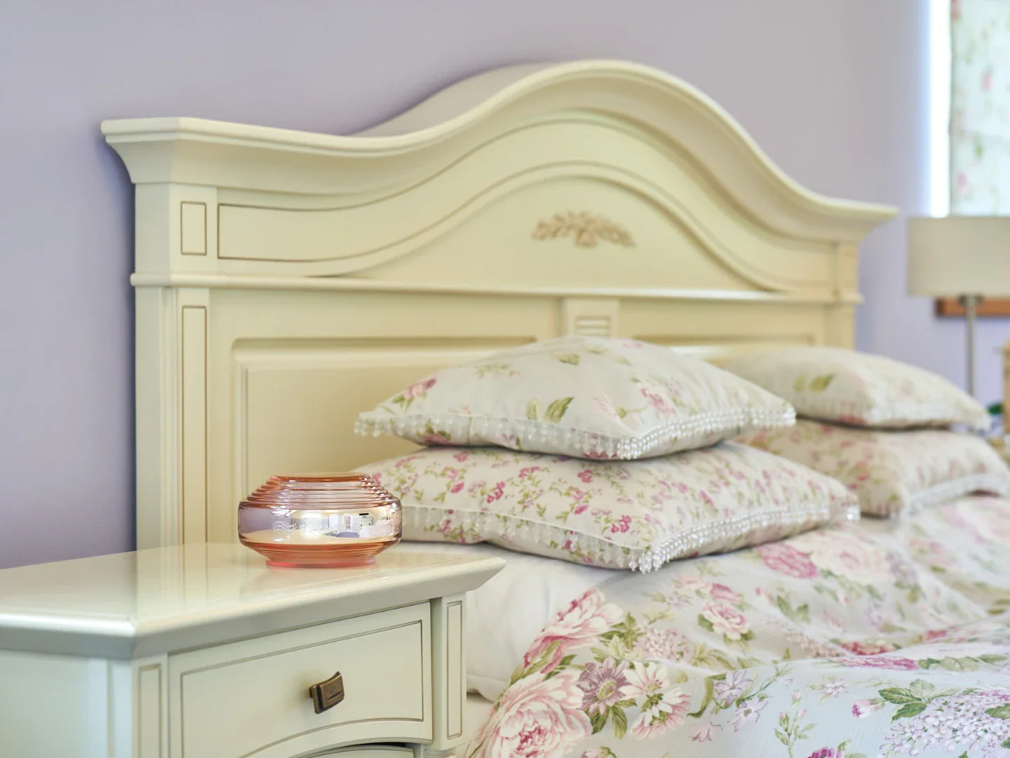 A bedroom featuring the Rosalin by Somavedic USA—a cream headboard, floral bedding, plush pillows, and a glass candle holder on a matching nightstand—creates an atmosphere ideal for supporting circadian rhythms.