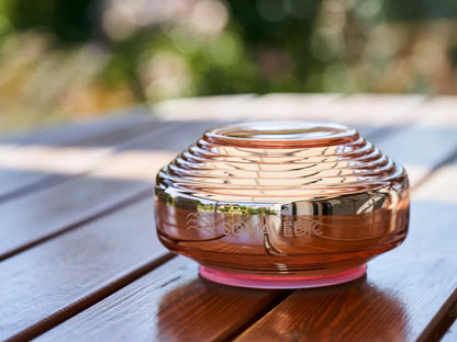 The Somavedic USA Rosalin, a glass device with a ribbed circular design, rests on a wooden outdoor table reflecting light—ideal for supporting circadian rhythm—set against a backdrop of blurred greenery.
