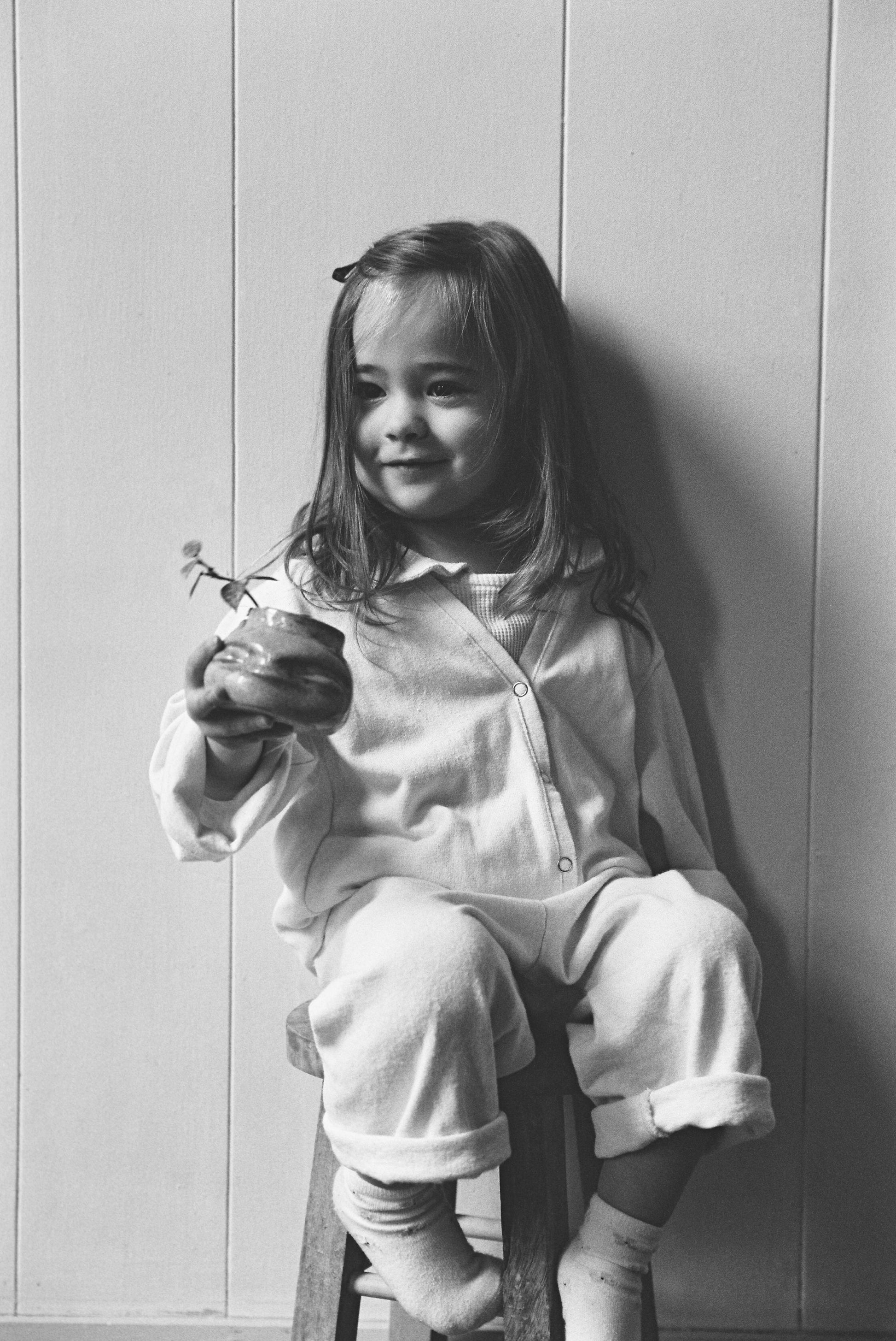 A young girl with long hair sits on a wooden stool, smiling and holding a small potted plant. She wears the cabane childrenswear TAKE CARE TOP hemp pajamas, paired with socks. The background is a plain, light-colored wall.