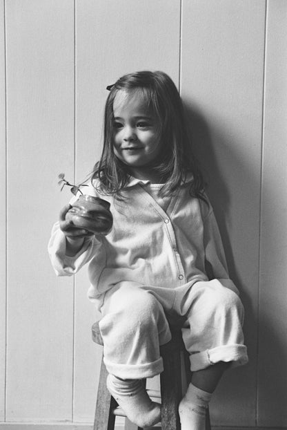 A young girl with long hair sits on a wooden stool, smiling and holding a small potted plant. She wears the cabane childrenswear TAKE CARE TOP hemp pajamas, paired with socks. The background is a plain, light-colored wall.