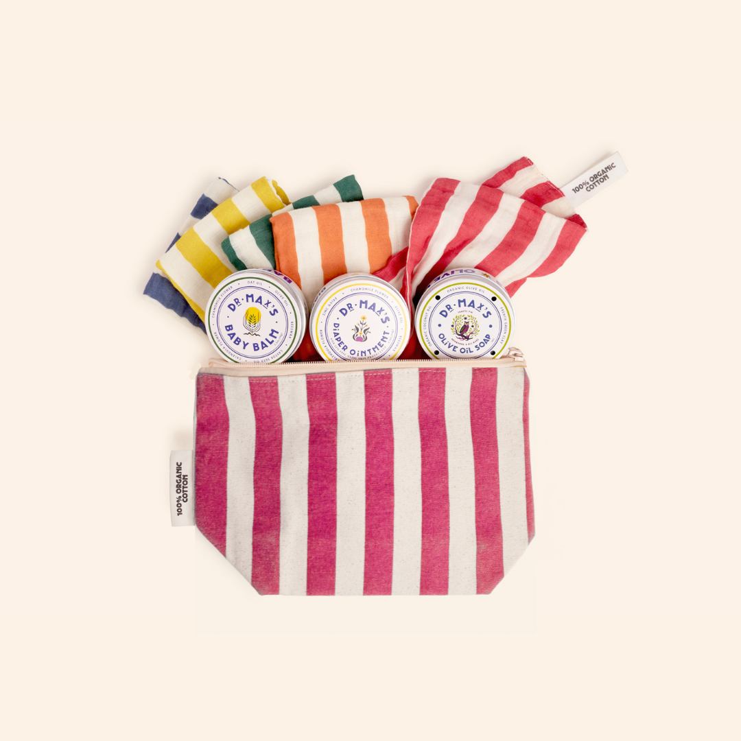 Dr. Max's Newborn Gift Box includes a pink and white striped pouch with two tins—Baby Balm and Baby Ointment—and several colorful cloths, all essentials for gentle newborn care, neatly arranged against a neutral background.