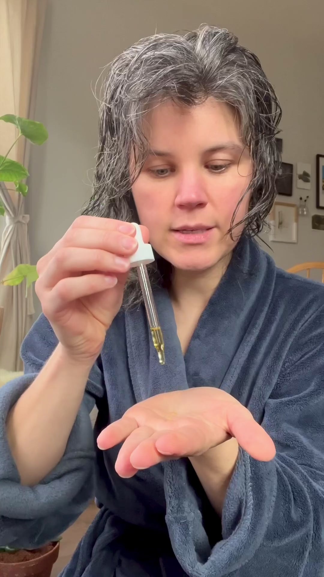 A person with wet hair in a blue robe applies & Tread Well Hair Oil from a dropper onto their palm indoors, near a window with plants and framed pictures, to repair damage and support hair growth.