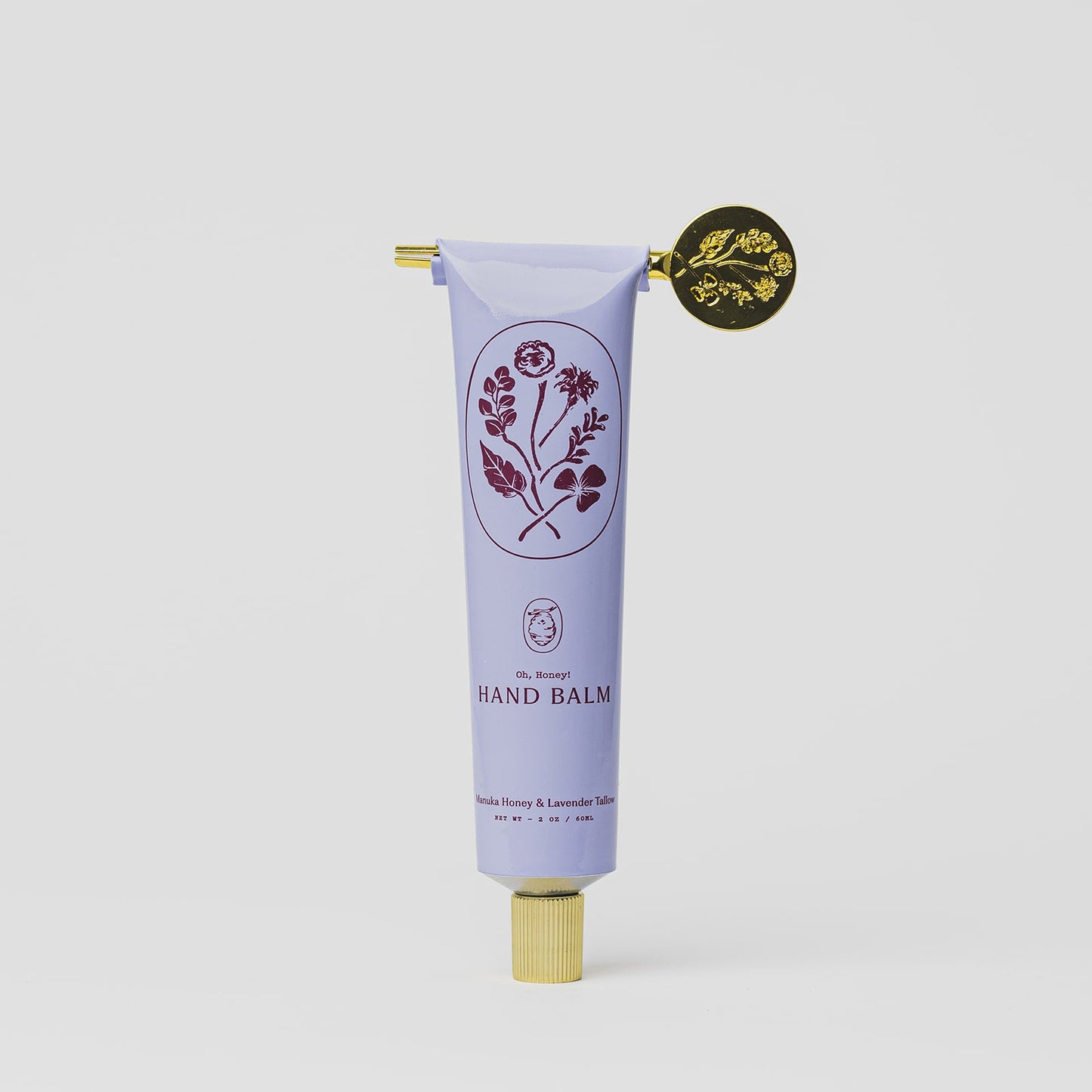 A lavender tube of My Neighbor’s Tallow Manuka Honey & Lavender hand balm with a gold cap, decorative gold seal, and floral illustrations, labeled “Hand Balm.” Scented with vanilla, honey, and lavender on a plain white background.