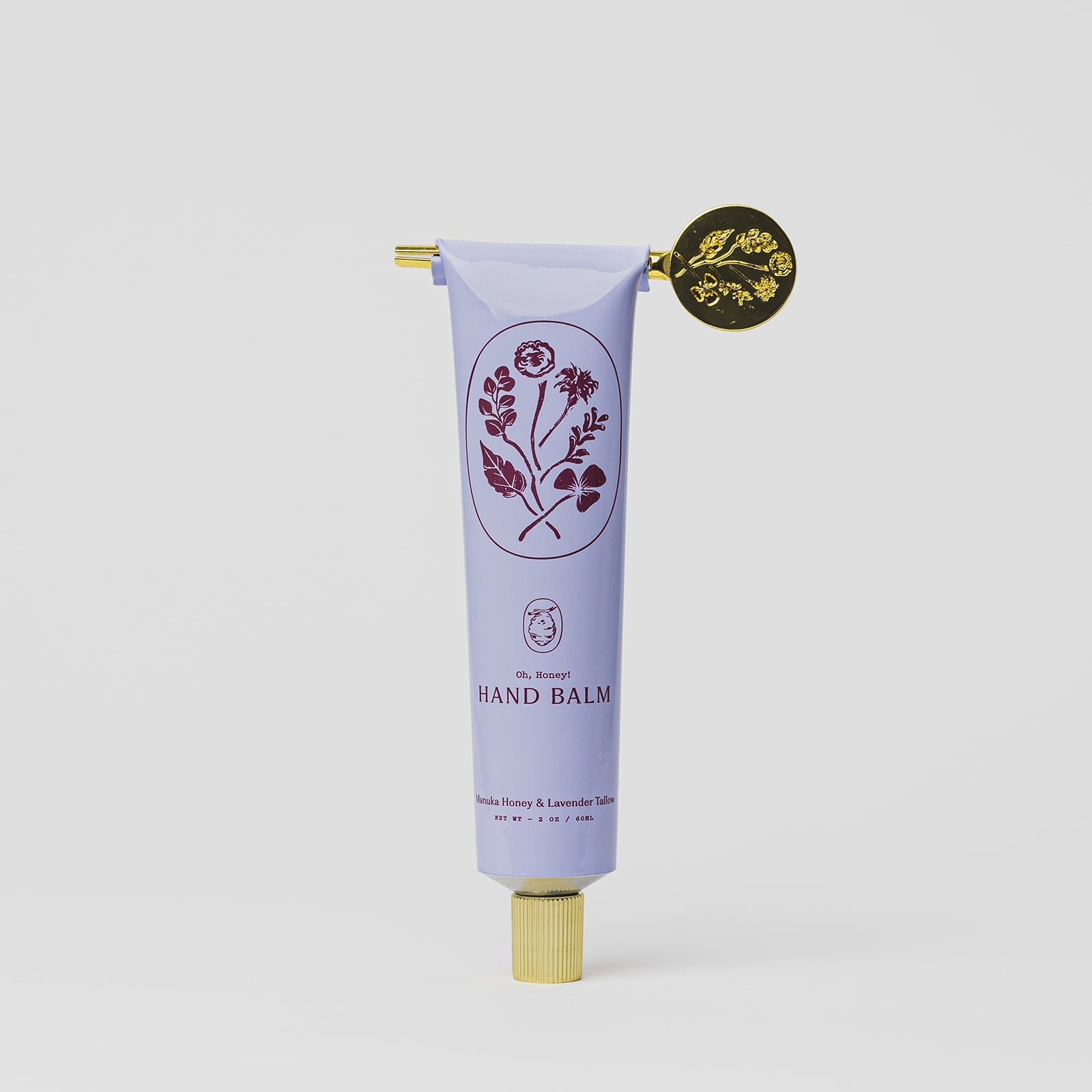 A lavender tube of My Neighbor’s Tallow Manuka Honey & Lavender hand balm with a gold cap, decorative gold seal, and floral illustrations, labeled “Hand Balm.” Scented with vanilla, honey, and lavender on a plain white background.