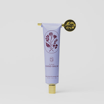 A lavender tube of My Neighbor’s Tallow Manuka Honey & Lavender hand balm with a gold cap, decorative gold seal, and floral illustrations, labeled “Hand Balm.” Scented with vanilla, honey, and lavender on a plain white background.
