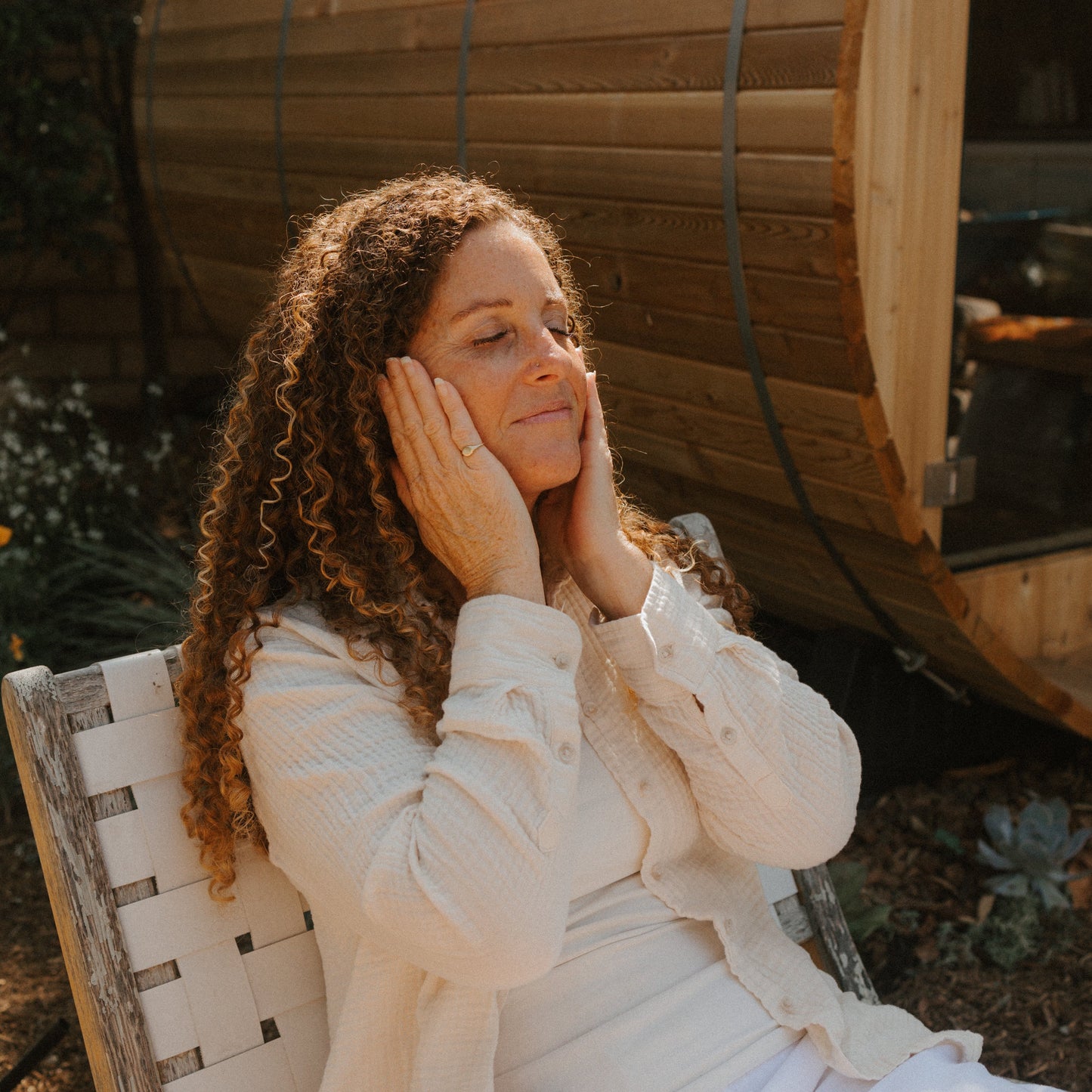 A woman with curly hair sits outdoors in a white outfit, eyes closed and hands on her cheeks, looking relaxed—perhaps enjoying the benefits of Honeybee Hippie’s antioxidant-rich saffron serum. A wooden sauna stands in the background.