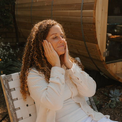A woman with curly hair sits outdoors in a white outfit, eyes closed and hands on her cheeks, looking relaxed—perhaps enjoying the benefits of Honeybee Hippie’s antioxidant-rich saffron serum. A wooden sauna stands in the background.