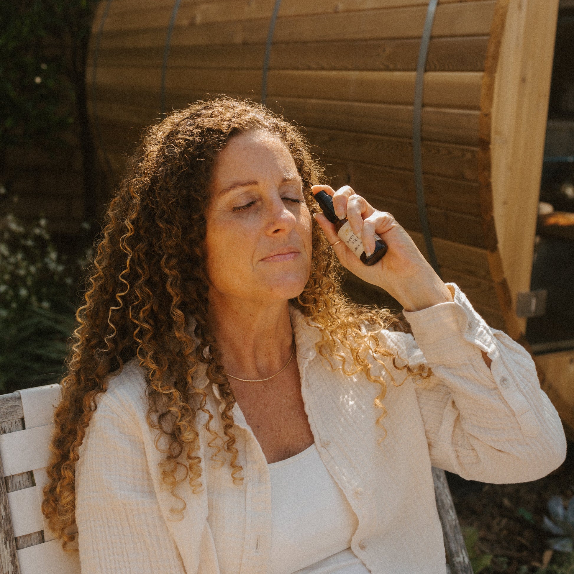 A woman with curly hair enjoys a refreshing moment outdoors, eyes closed as she sprays Honeybee Hippie's saffron serum onto her face, embracing antioxidant-rich skincare in front of a wooden structure.