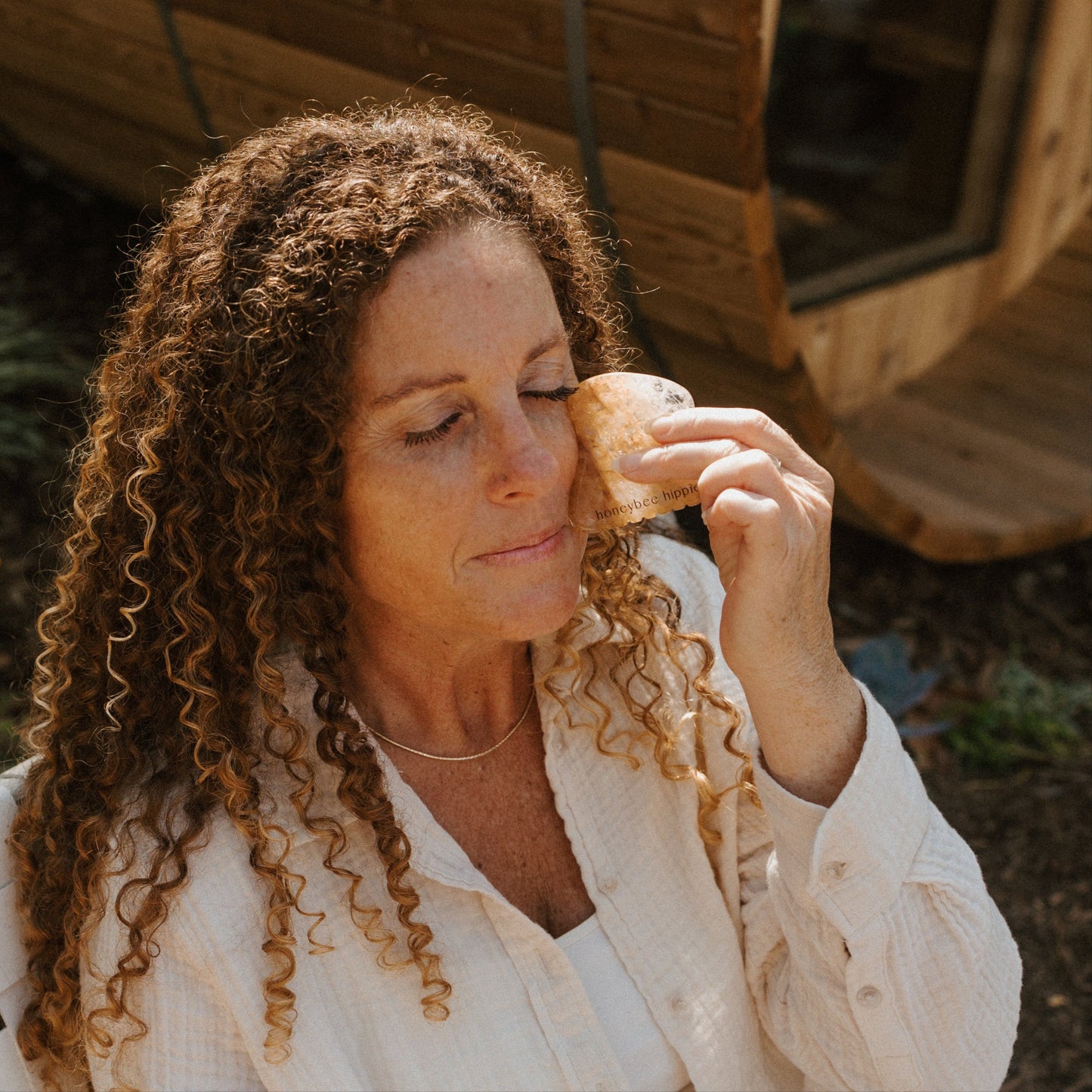 A woman with curly hair and a white shirt sits outdoors, gently using the Honeybee Hippie golden quartz gua sha on her closed eye, looking calm and relaxed.