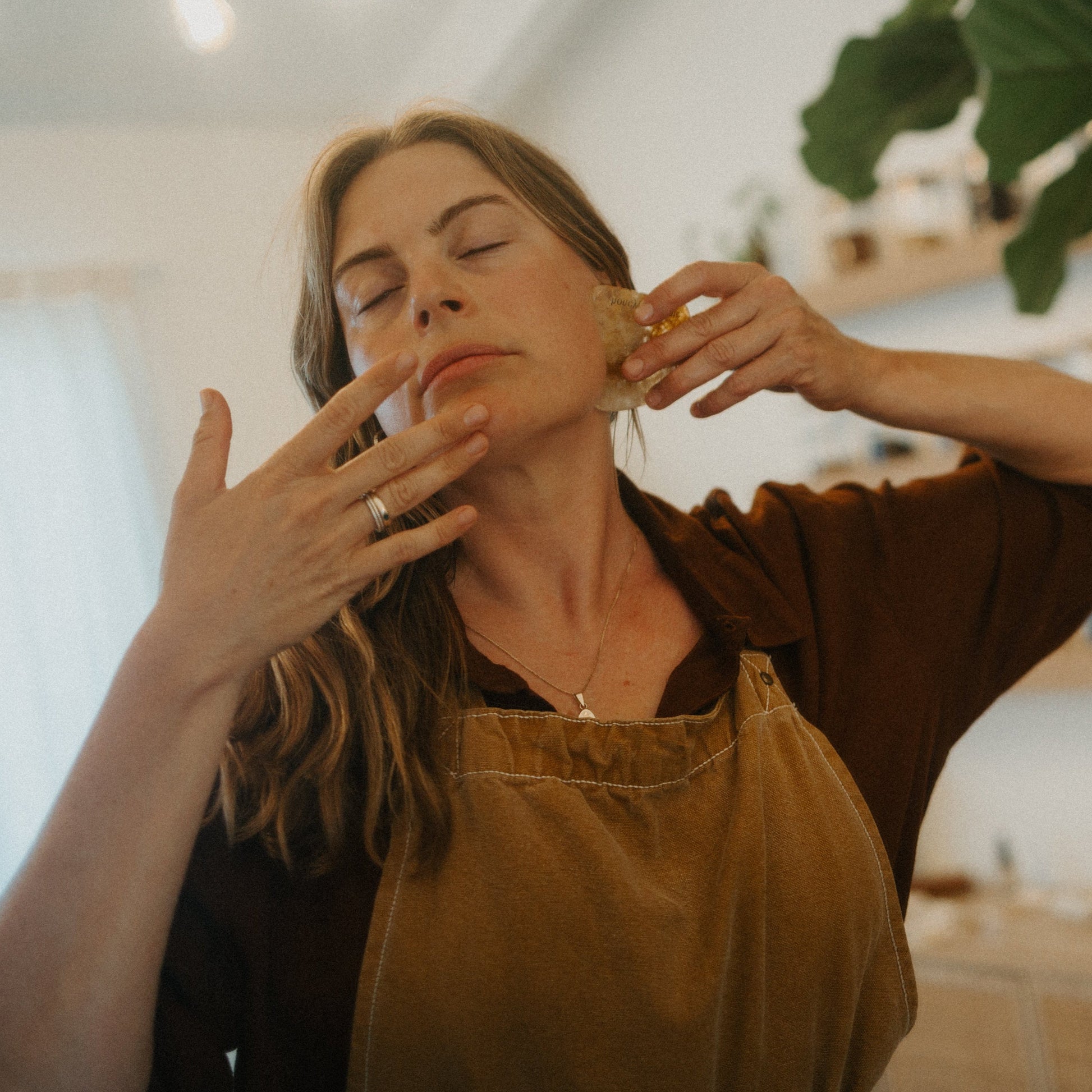 A woman in a brown apron gently massages her jawline with the Honeybee Hippie golden quartz gua sha, eyes closed in relaxation, standing indoors under soft lighting for a soothing lymphatic drainage ritual.
