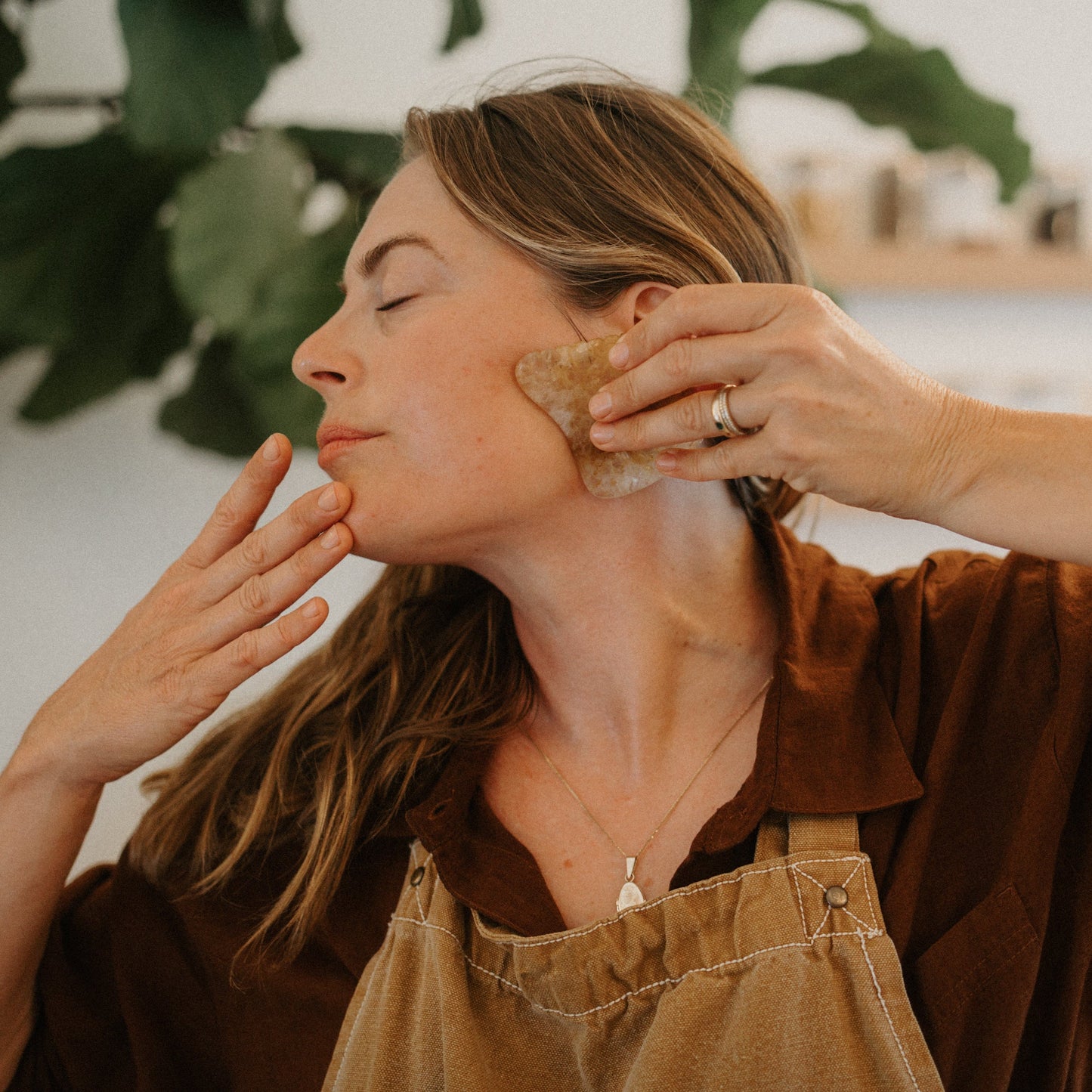 A woman with long hair uses the Honeybee Hippie golden quartz gua sha on her jawline, eyes closed in relaxation. Wearing a brown shirt and apron, she enjoys lymphatic drainage as green leaves and shelves blur softly behind her.