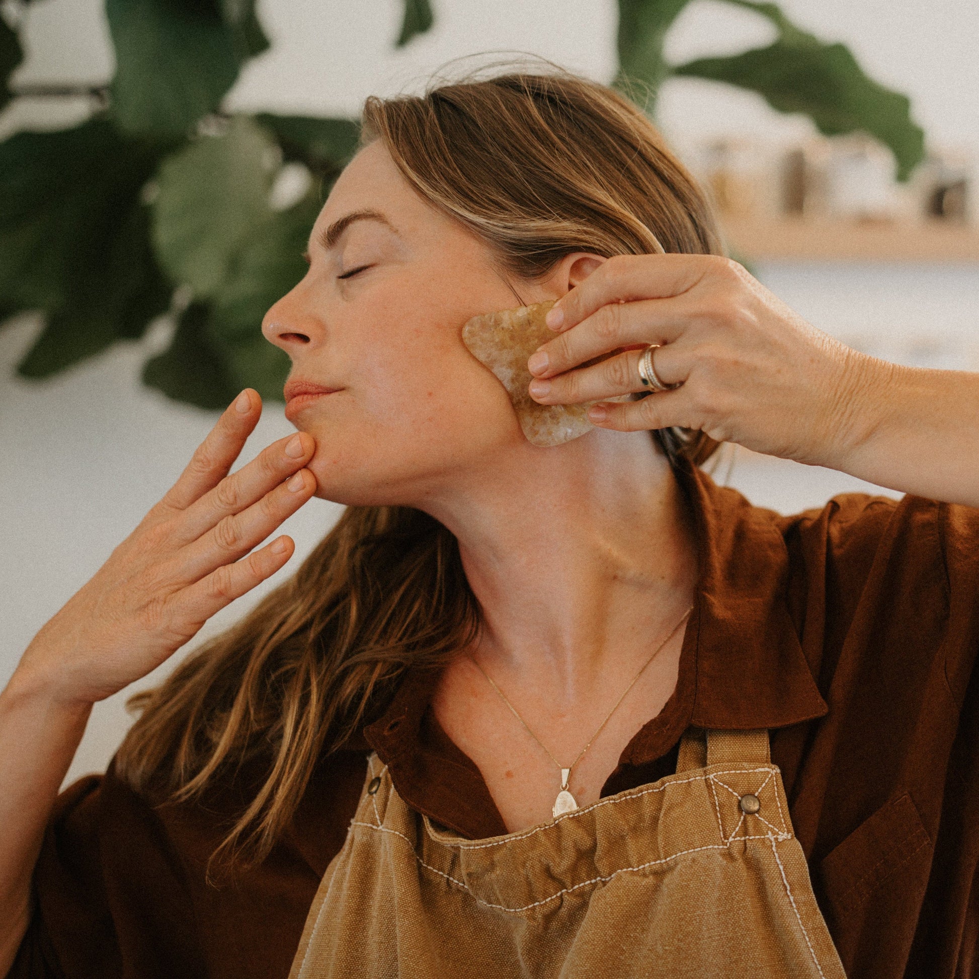 A woman with long hair uses the Honeybee Hippie golden quartz gua sha on her jawline, eyes closed in relaxation. Wearing a brown shirt and apron, she enjoys lymphatic drainage as green leaves and shelves blur softly behind her.