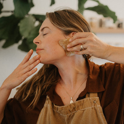 A woman with long hair uses the Honeybee Hippie golden quartz gua sha on her jawline, eyes closed in relaxation. Wearing a brown shirt and apron, she enjoys lymphatic drainage as green leaves and shelves blur softly behind her.