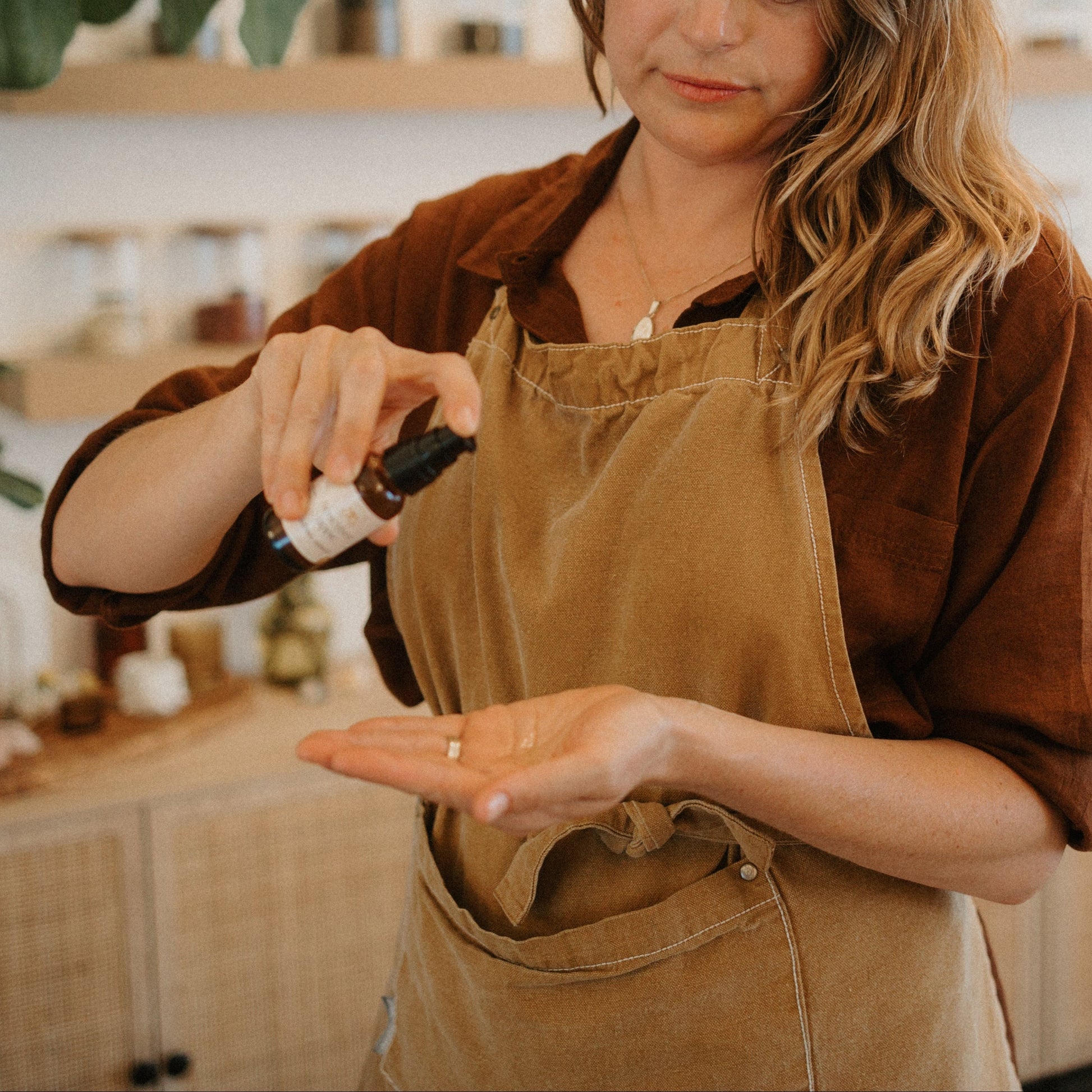 In a cozy, softly lit room, a woman in a brown shirt and tan apron dispenses Honeybee Hippie saffron serum, known for its antioxidants, from a small dropper bottle onto her hand.