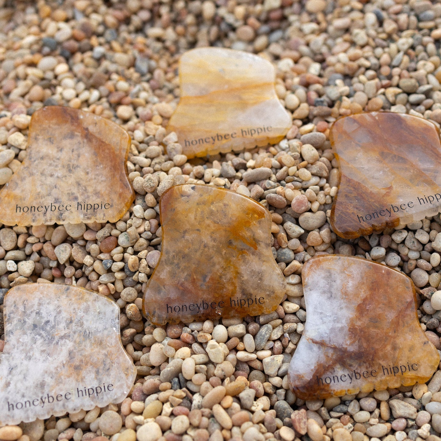 Six polished, irregular golden quartz gua sha tools by Honeybee Hippie are displayed on pebbles. Each brown, yellow, and cream stone is engraved with the brand name—ideal for facial sculpting and lymphatic drainage.