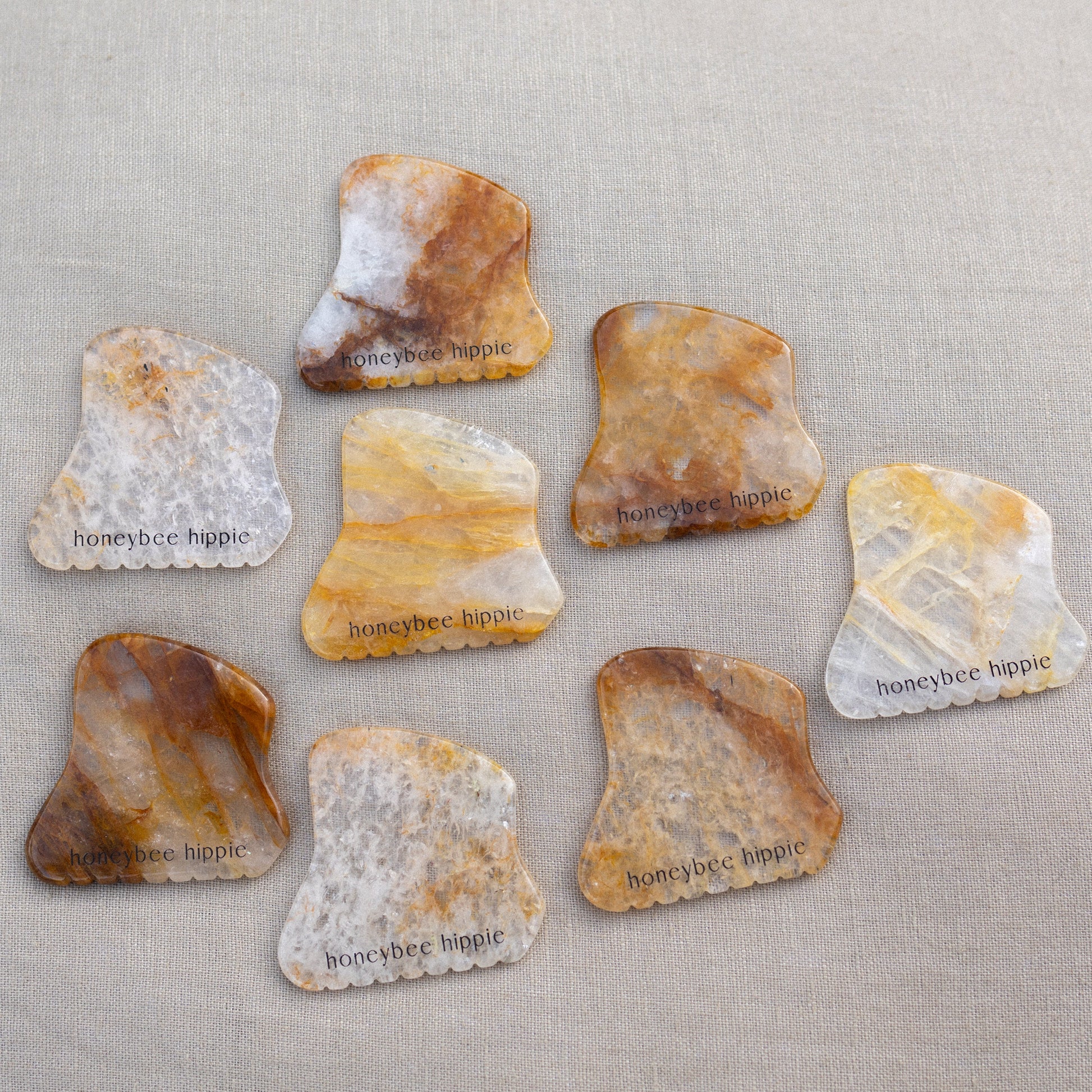 Eight polished, irregular Golden Quartz Gua Sha stones from Honeybee Hippie, each engraved with "honeybee hippie," display natural brown, yellow, and gray patterns on a neutral fabric background.