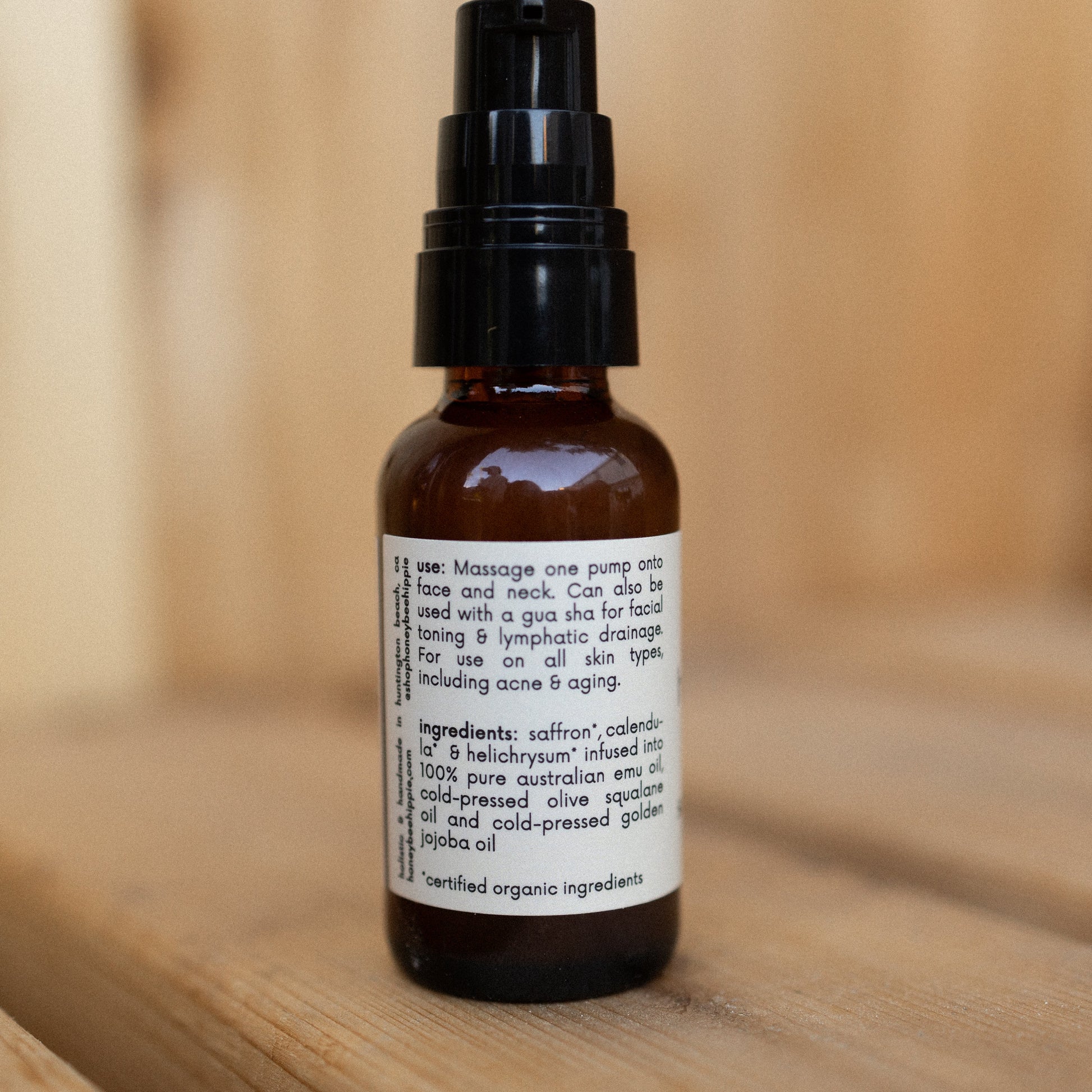 A brown glass bottle with a black pump top, labeled with directions and ingredients for Honeybee Hippie’s antioxidant-rich saffron serum, is displayed against a wooden background.