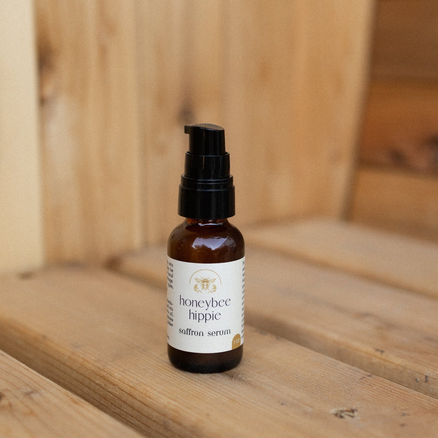A small amber glass bottle with a black pump labeled "Honeybee Hippie saffron serum" sits on wood, highlighting its antioxidant-rich formula designed to reduce hyperpigmentation, set against a softly blurred wooden background.