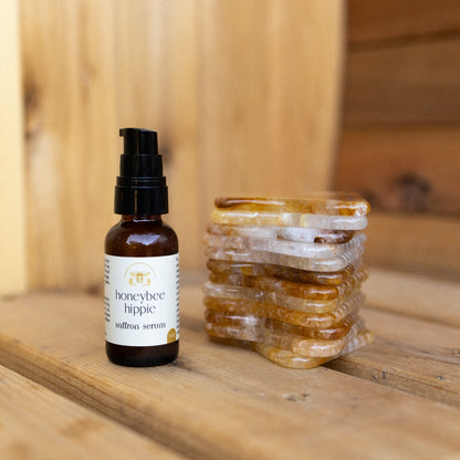 A bottle of Honeybee Hippie saffron serum rests on a wooden surface next to a stacked pile of stone gua sha tools in yellow, white, and amber tones.