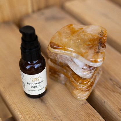 A small Honeybee Hippie Saffron Serum bottle—an antioxidant-rich blend for hyperpigmentation—stands next to orange and white stone gua sha tools on a wooden surface.
