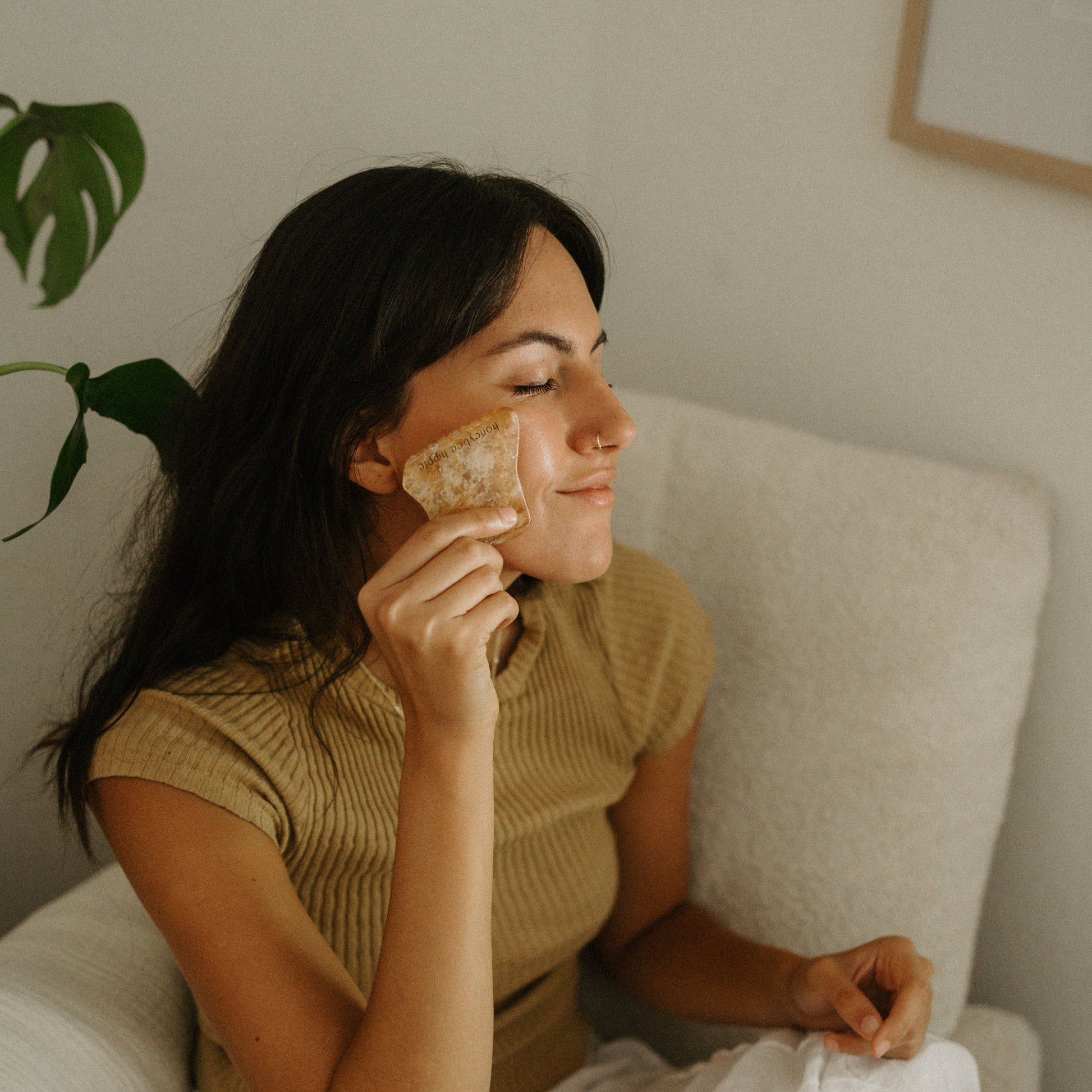 A woman with long dark hair relaxes in a cream chair, smiling with her eyes closed as she massages her cheek using the Honeybee Hippie golden quartz gua sha, enjoying a soothing facial ritual in a short-sleeved ribbed top.