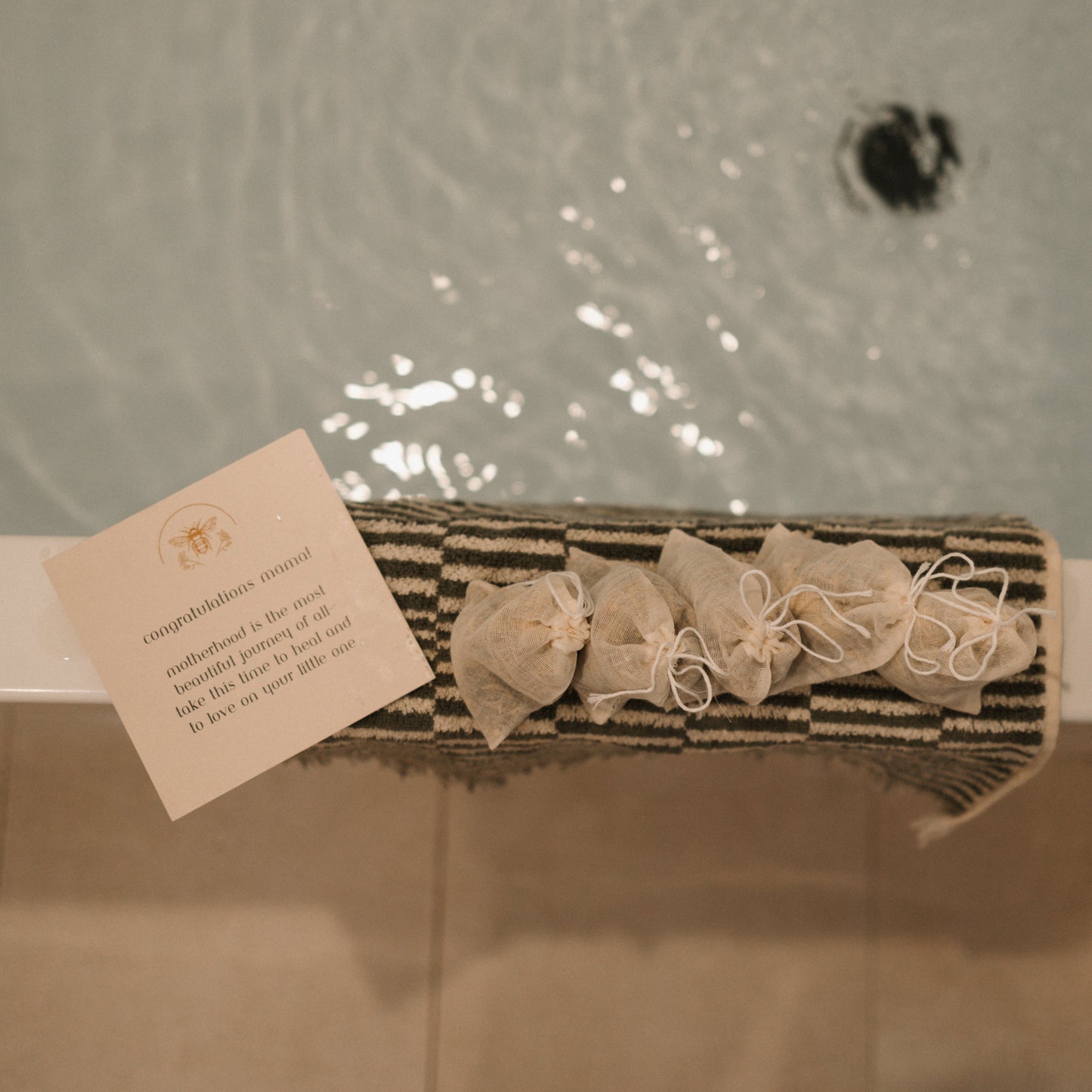 A note and small, tied pouches rest on a striped mat by the bathtub. It congratulates the new mom and suggests using Honeybee Hippie's postpartum herbal sitz bath for relaxation and perineal healing, perfect for comforting postpartum moms.
