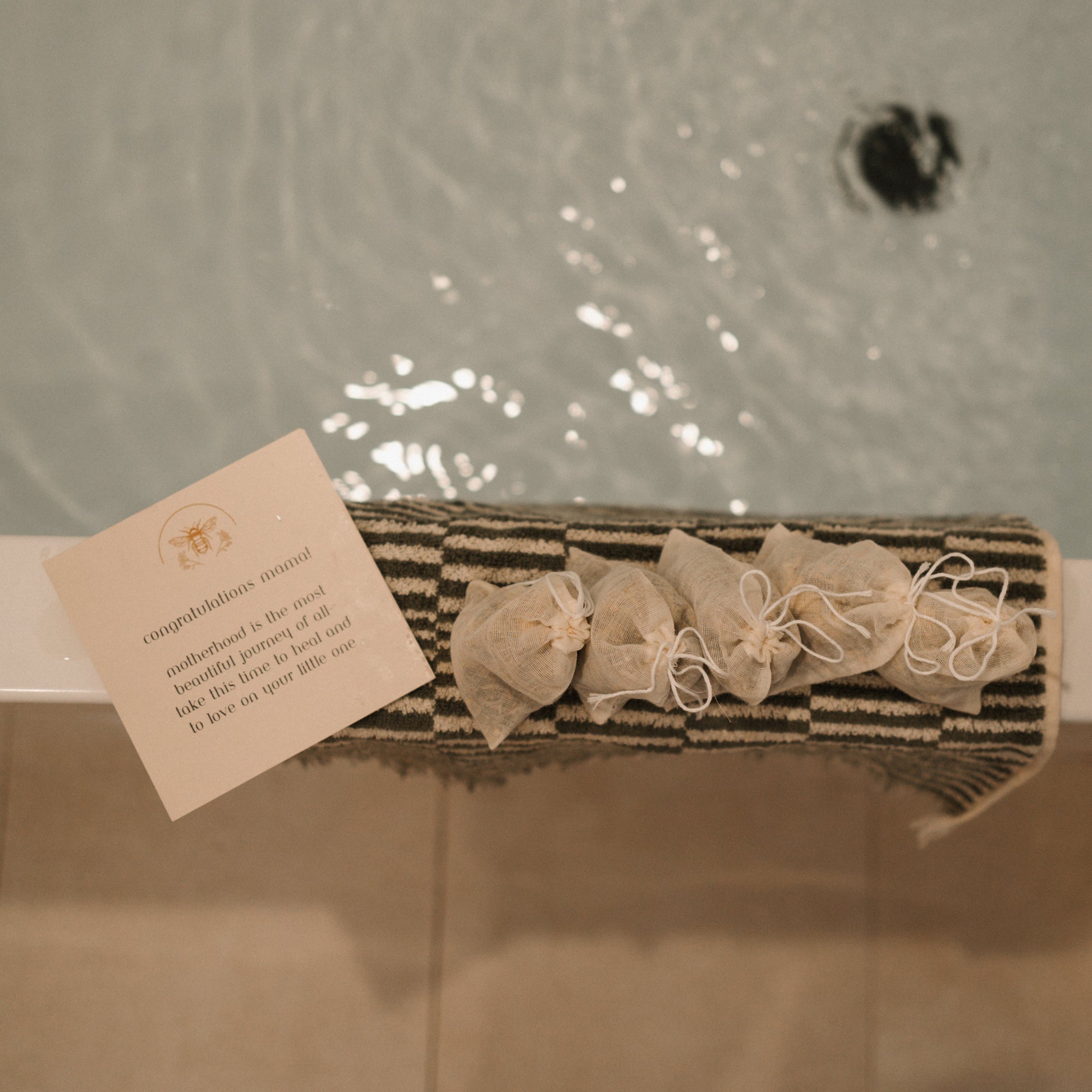 A note and small, tied pouches rest on a striped mat by the bathtub. It congratulates the new mom and suggests using Honeybee Hippie's postpartum herbal sitz bath for relaxation and perineal healing, perfect for comforting postpartum moms.