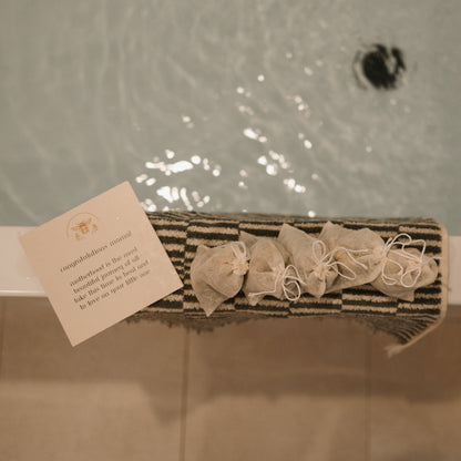 A note and small, tied pouches rest on a striped mat by the bathtub. It congratulates the new mom and suggests using Honeybee Hippie's postpartum herbal sitz bath for relaxation and perineal healing, perfect for comforting postpartum moms.