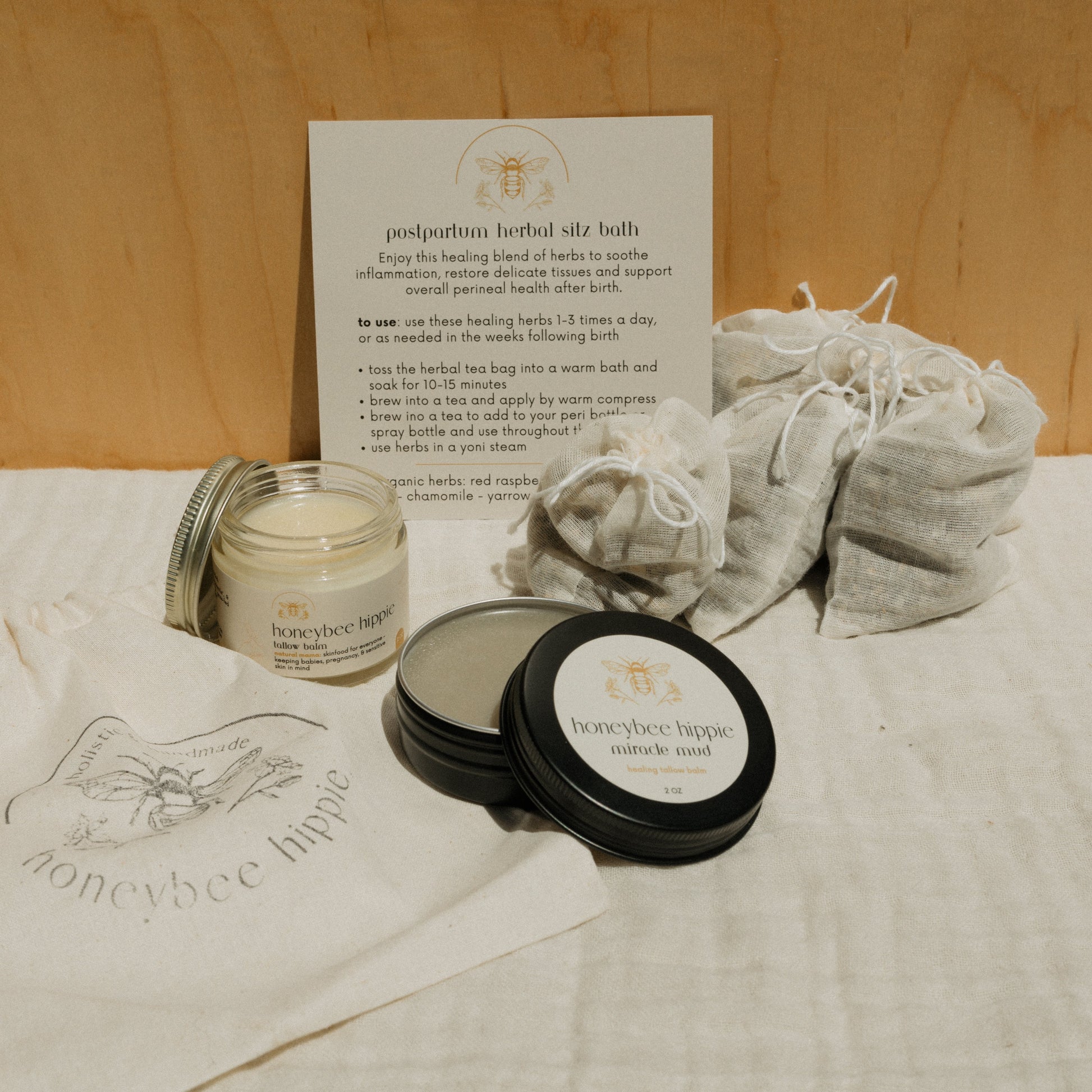 The Natural Mama Bundle by Honeybee Hippie features a postpartum herbal sitz bath kit, balm, miracle mud tin, herbal pouches, instruction card, and linen bag—all arranged on neutral fabric for a gentle self-care experience.