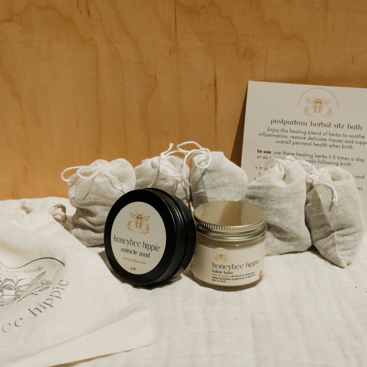The Honeybee Hippie natural mama bundle features jars of Natural Mama Tallow and Miracle Mud, small cloth pouches, and an instruction card for a postpartum herbal sitz bath, all arranged on a light fabric.