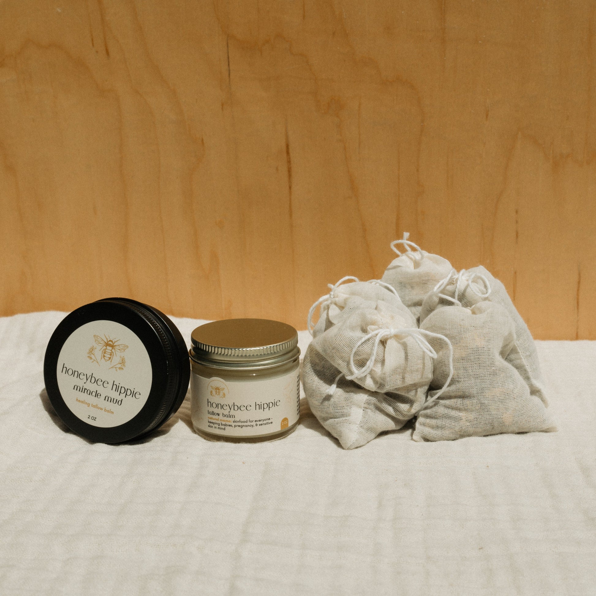 Jars labeled "honeybee hippie miracle mud" and "honeybee hippie body butter" sit on a cream cloth, next to small fabric pouches. The warm wooden background completes the Honeybee Hippie's "the natural mama bundle" vibe perfectly.