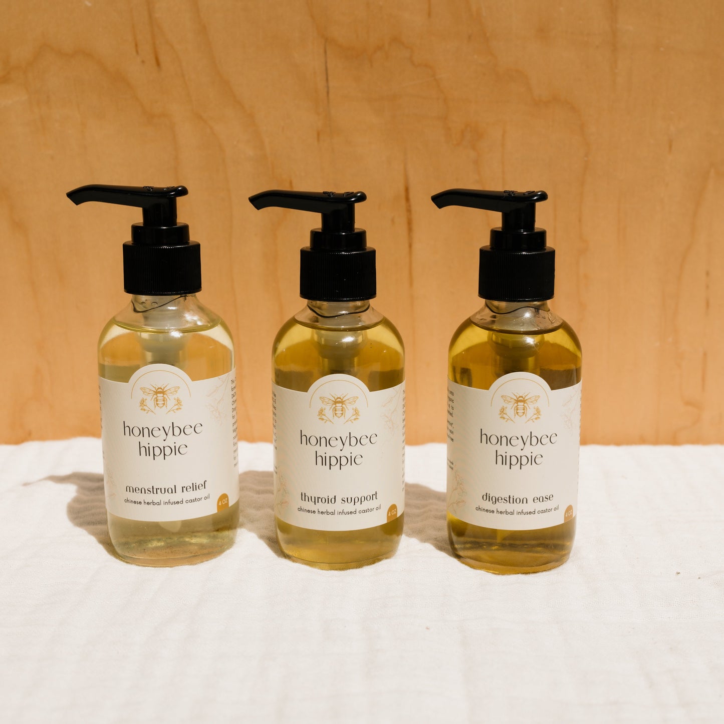 Three bottles of liquid products with pump dispensers labeled "Honeybee Hippie" are set against a wooden background. They include "menstrual relief," "thyroid support," and "digestion ease," all promoting holistic health with beneficial Chinese herbs.