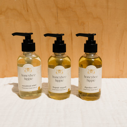 Three bottles of liquid products with pump dispensers labeled "Honeybee Hippie" are set against a wooden background. They include "menstrual relief," "thyroid support," and "digestion ease," all promoting holistic health with beneficial Chinese herbs.