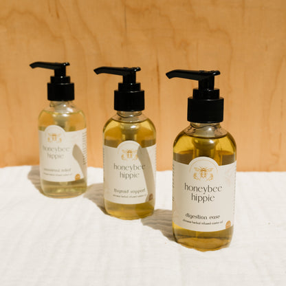 Three bottles of Honeybee Hippie's herbal infused castor oil packs, enriched with Chinese herbs, sit on a white surface against a wooden background. The labels claim to aid digestion, support the thyroid, and provide emotional relief, all featuring black pump dispensers for holistic benefits.