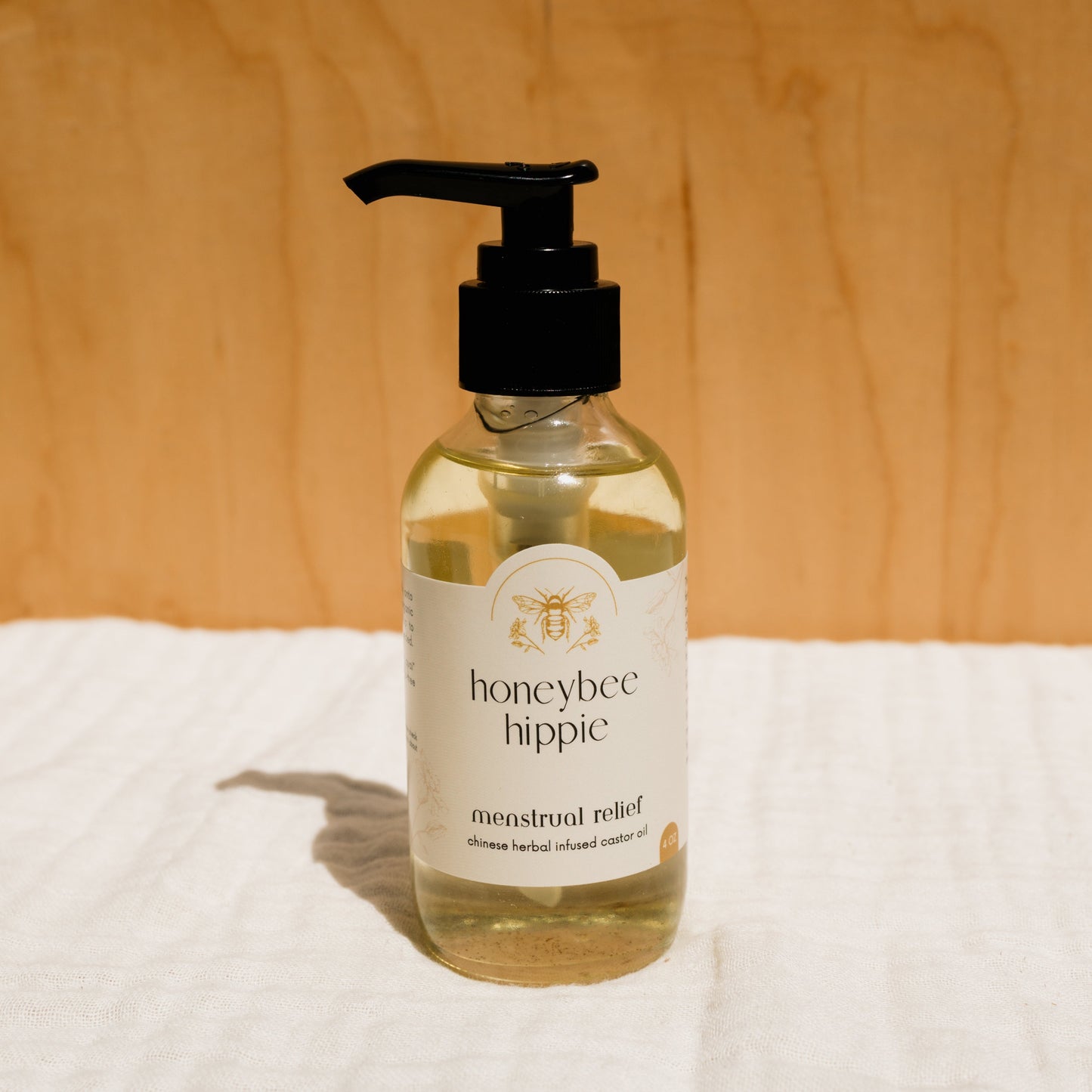 A clear pump bottle labeled "Honeybee Hippie" featuring "menstrual relief" sits on a white cloth with a wooden backdrop. It contains light-colored herbal infused castor oil packs, embodying nature's holistic comfort.