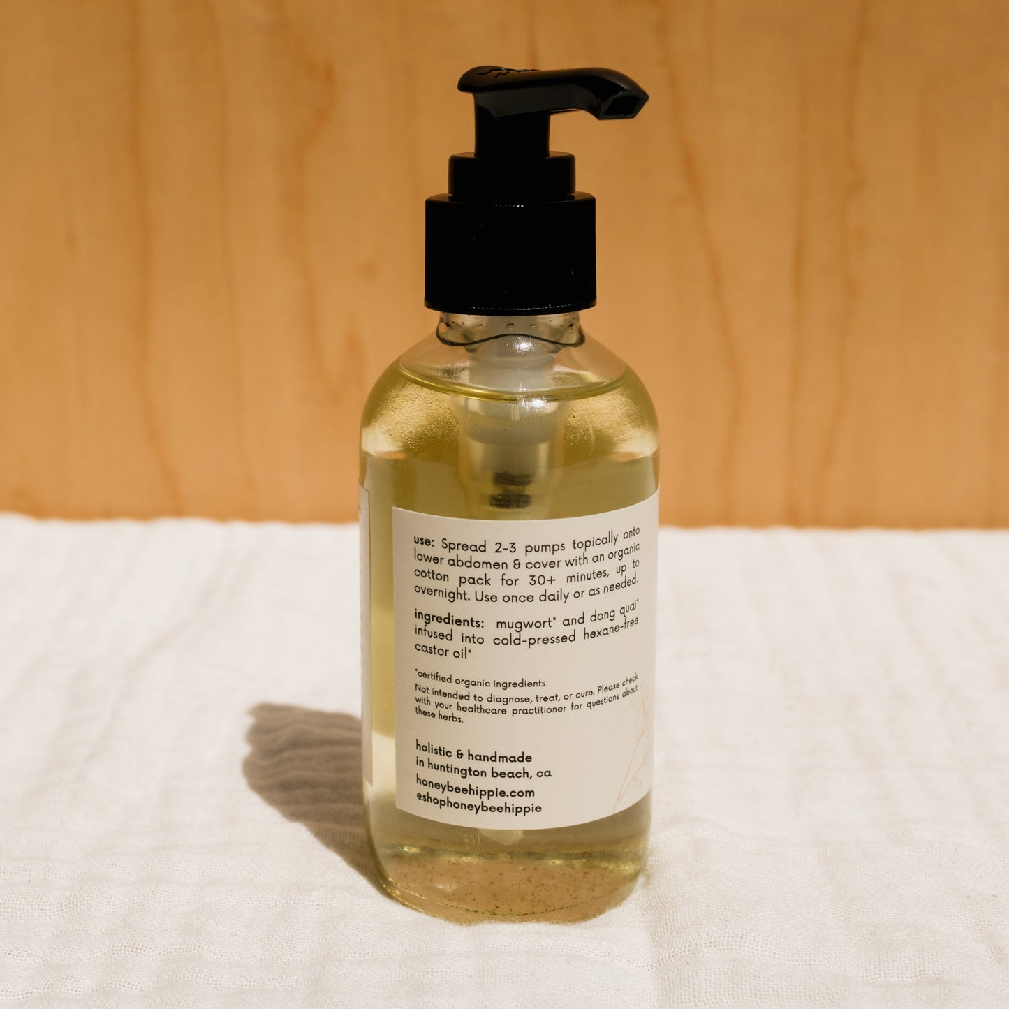 A clear pump bottle containing herbal-infused castor oil by Honeybee Hippie sits on a white cloth. The label highlights its blend of castor oil and Chinese herbs, with instructions and ingredients, all set against a wooden background that enhances its holistic health appeal.
