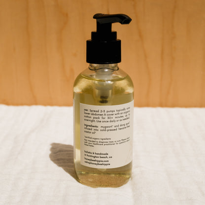 A clear pump bottle containing herbal-infused castor oil by Honeybee Hippie sits on a white cloth. The label highlights its blend of castor oil and Chinese herbs, with instructions and ingredients, all set against a wooden background that enhances its holistic health appeal.