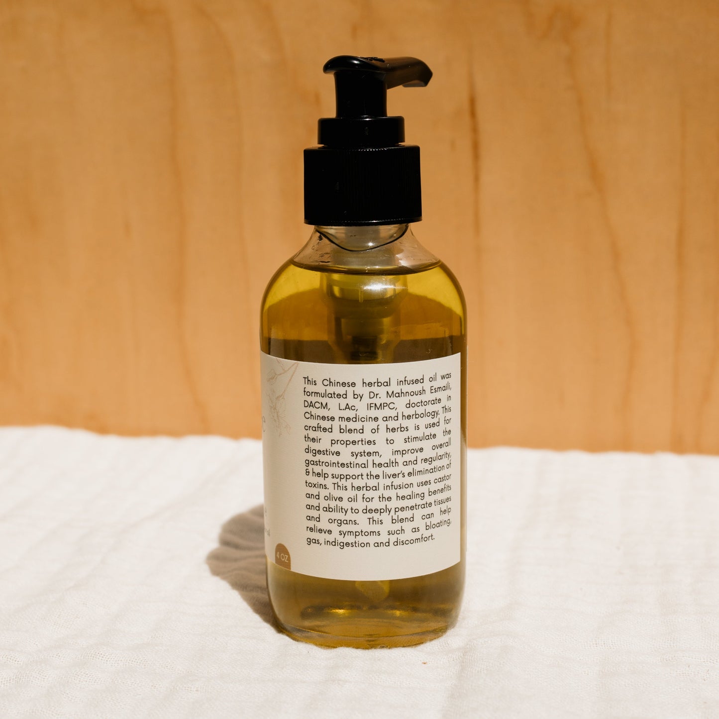 A clear pump bottle of Honeybee Hippie's herbal-infused castor oil packs sits against a wooden backdrop. The label highlights its Chinese herbal formula, promoting digestion and wellness while emphasizing soothing properties for holistic health.