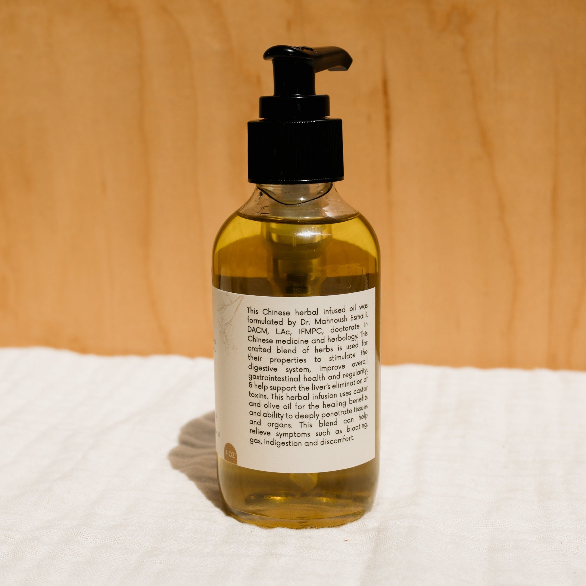 A clear pump bottle of Honeybee Hippie's herbal-infused castor oil packs sits against a wooden backdrop. The label highlights its Chinese herbal formula, promoting digestion and wellness while emphasizing soothing properties for holistic health.