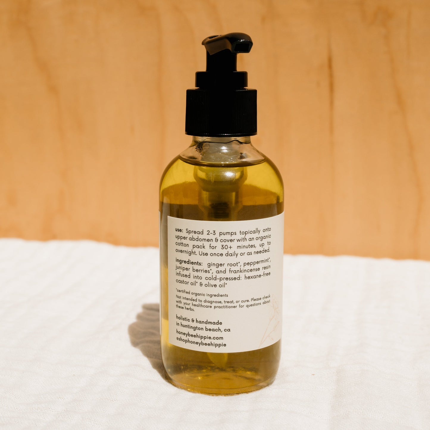A Honeybee Hippie herbal-infused castor oil pack with a pump dispenser sits against a wooden background. Featuring ingredients like ginger root, peppermint, and castor oil with usage instructions, it's ideal for those embracing holistic health practices.