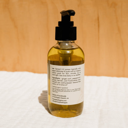 A Honeybee Hippie herbal-infused castor oil pack with a pump dispenser sits against a wooden background. Featuring ingredients like ginger root, peppermint, and castor oil with usage instructions, it's ideal for those embracing holistic health practices.