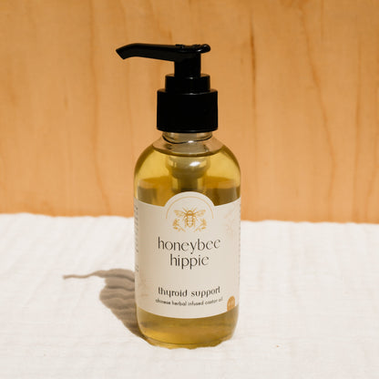 A bottle of Honeybee Hippie herbal infused castor oil packs with a pump dispenser sits on a white cloth, displaying its bee-illustrated label and "Chinese Herbal Infused Castor Oil" text, embracing holistic health against a wooden background.