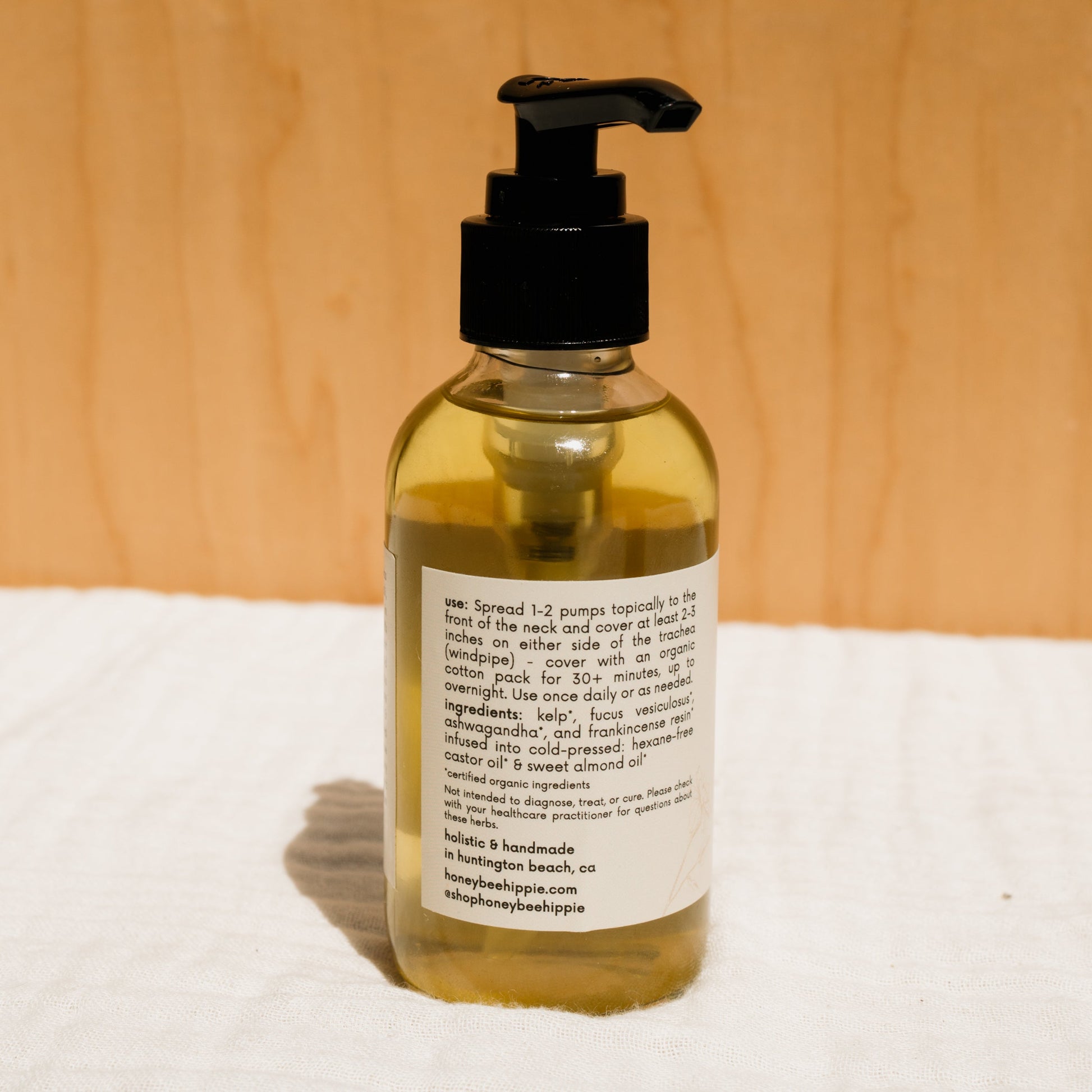 The Honeybee Hippie's herbal infused castor oil packs come in a bottle with a black pump top. The back label provides instructions, ingredients such as castor oil, and company details. Set against a light wood surface, sunlight casts a shadow that enhances its holistic health appeal.