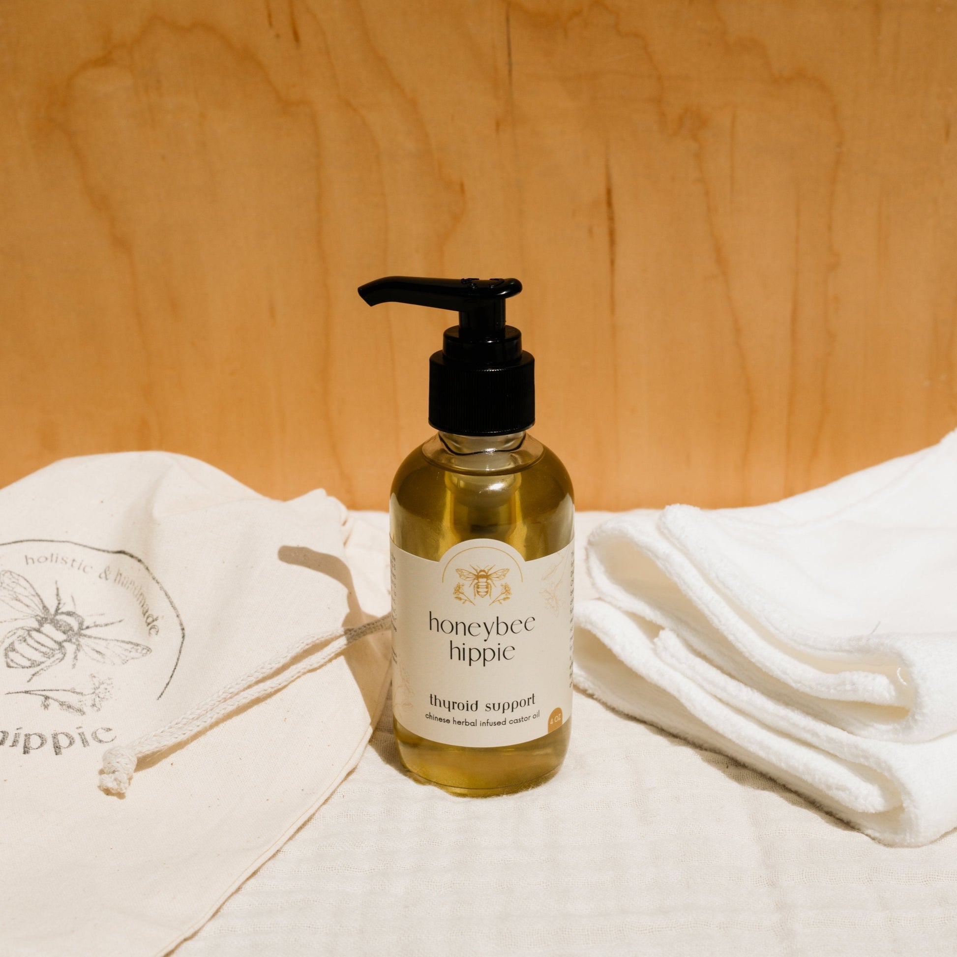 A bottle of Honeybee Hippie's "herbal infused castor oil packs" with a pump sits on a soft surface, surrounded by white cloths and a drawstring bag displaying a bee logo, set against a light wooden backdrop.