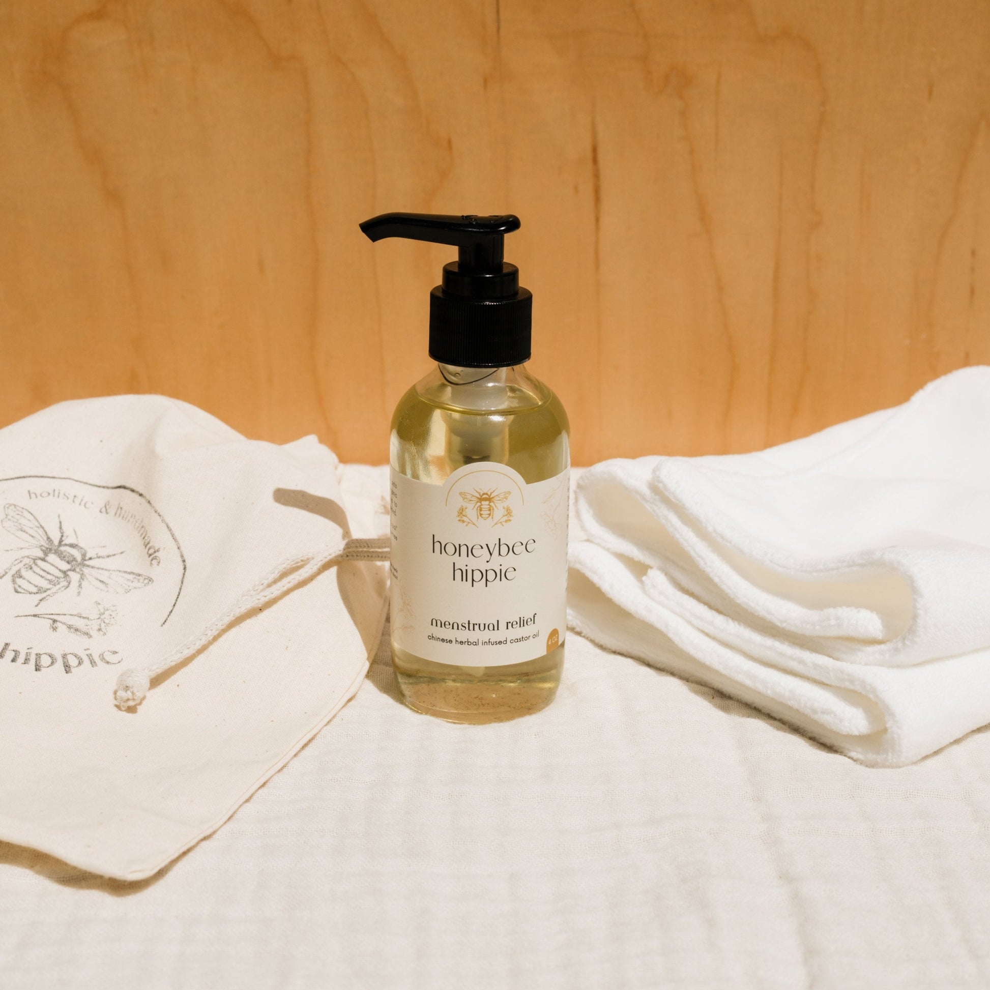 A bottle of Honeybee Hippie herbal infused castor oil packs with a pump dispenser is nestled between a cloth bag with a bee logo and folded white towels, embracing holistic health. A wooden wall and white fabric surface in the background enhance the blend of serenity and nature.