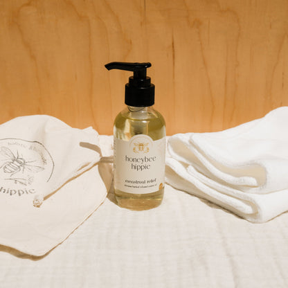 A bottle of Honeybee Hippie herbal infused castor oil packs with a pump dispenser is nestled between a cloth bag with a bee logo and folded white towels, embracing holistic health. A wooden wall and white fabric surface in the background enhance the blend of serenity and nature.